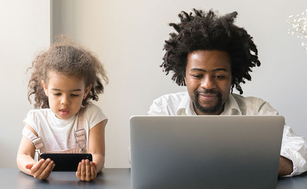 Photo of father and daughter looking at a laptop and a phone.
