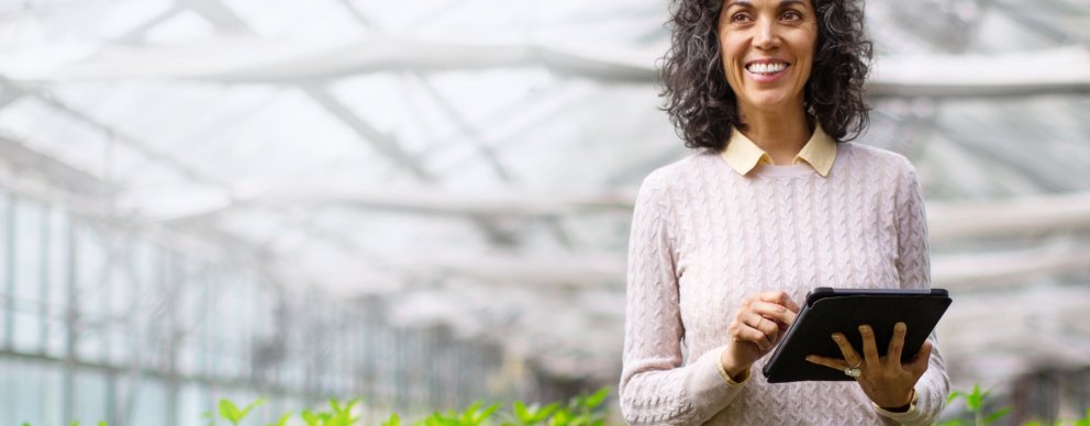 Woman standing in a greenhouse holding a tablet and smiling